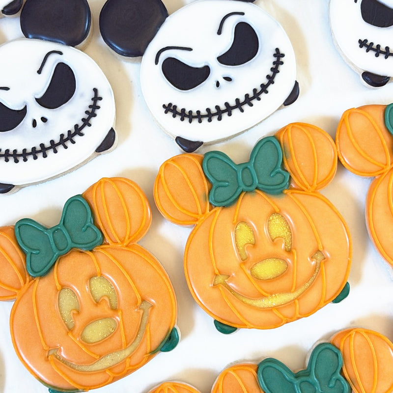 Halloween sugar cookies: grinning skeleton faces with round black ears and orange jack-o'-lanterns with teal bows